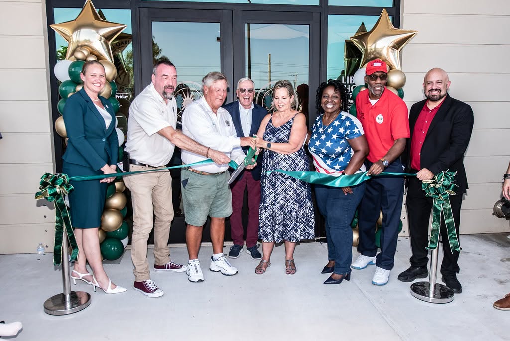Group of diverse adults at a ribbon-cutting ceremony outside a storefront, cutting a teal ribbon with balloons in the background.