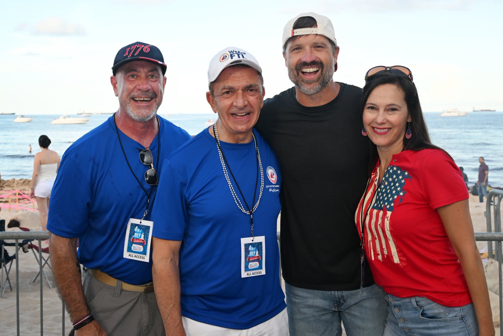 Group of four smiling adults posing on a beach, with the ocean and boats in the background.