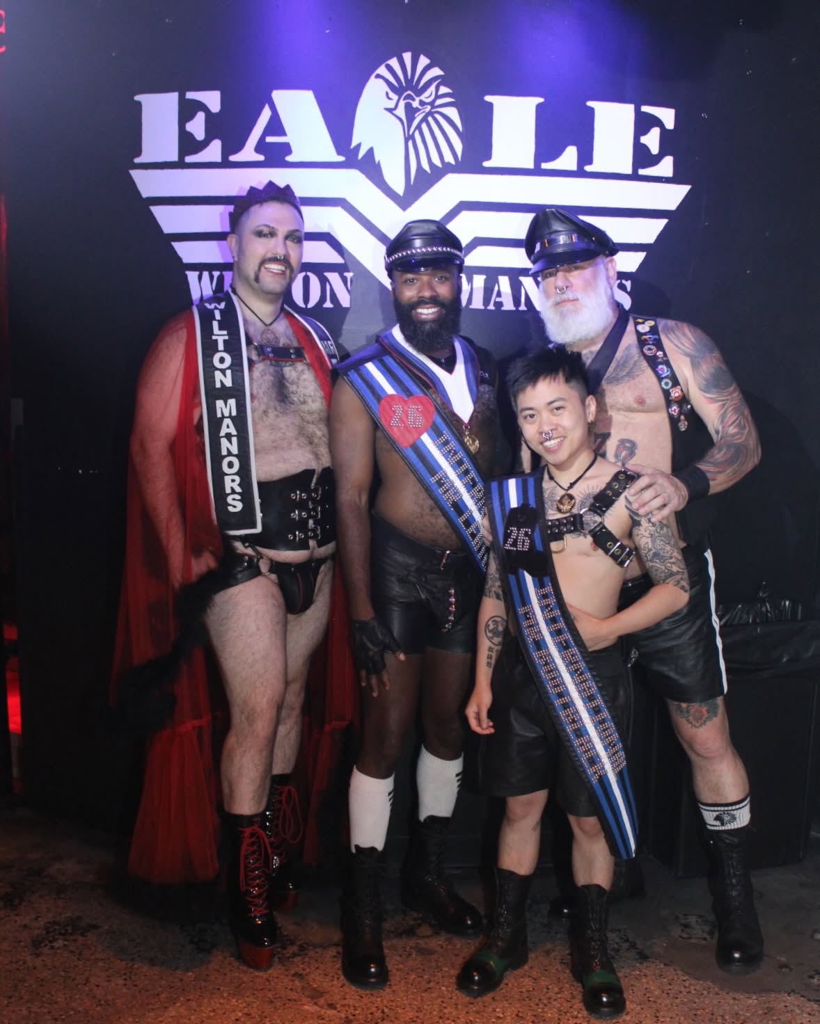 Group of four people in leather attire posing together in front of a large Eagle logo backdrop at a leather event.