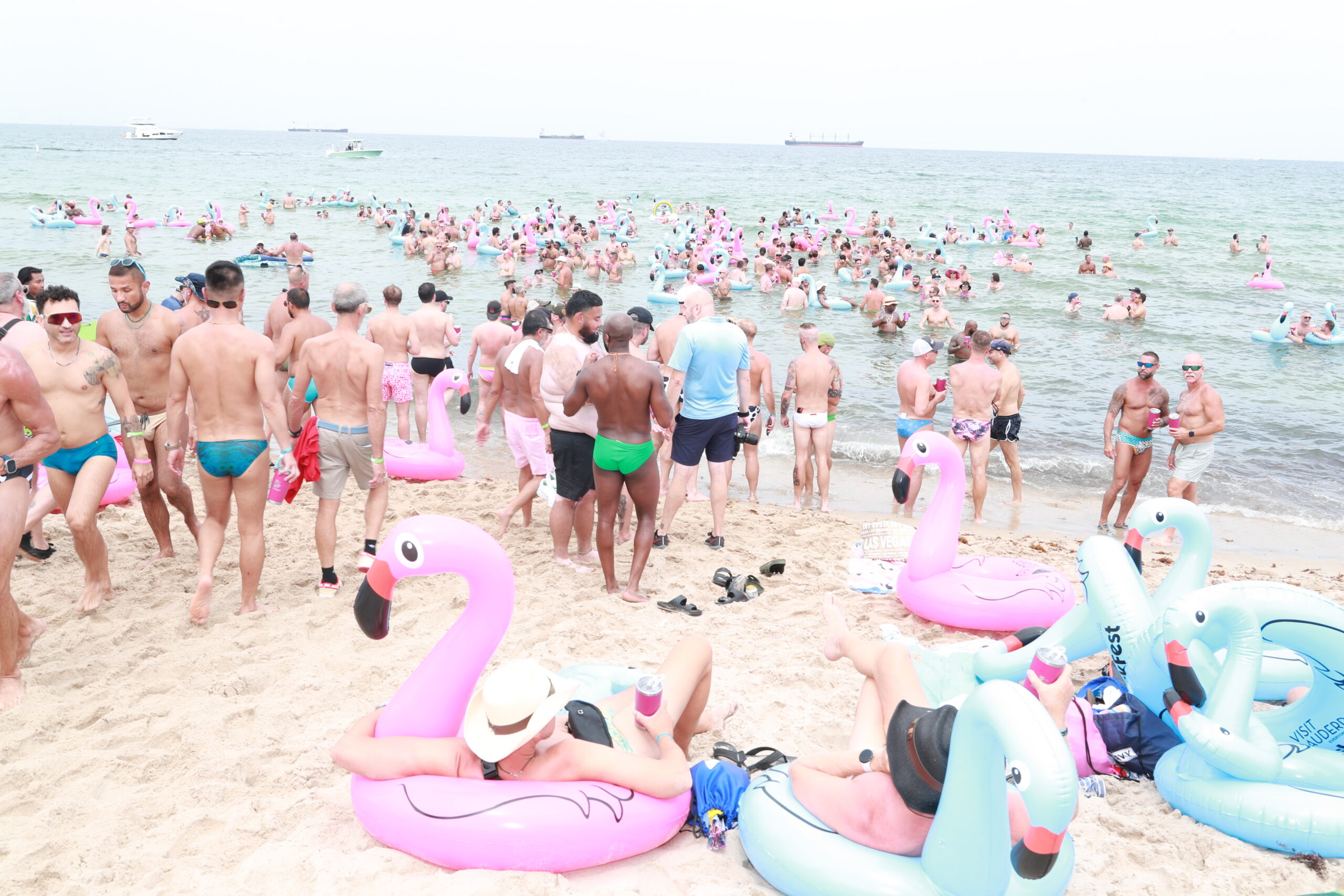 Crowded beach scene with people in swimsuits and large pink flamingo inflatables along the shore and in the water.