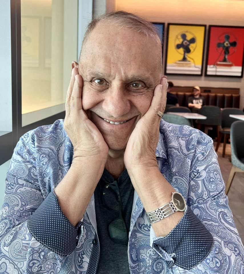 Smiling older man rests his face in his hands at a cafe, wearing paisley-patterned shirt and a silver watch.