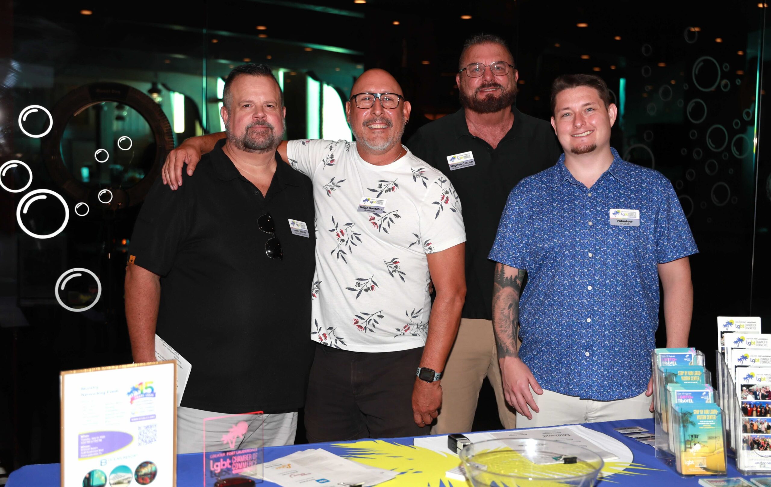 Out With Scott – By Scott Holland | Hotspots Magazine Hotspots Magazine Four men stand together at an event booth, smiling, with LGBT travel brochures and pamphlets on the table in front of them.