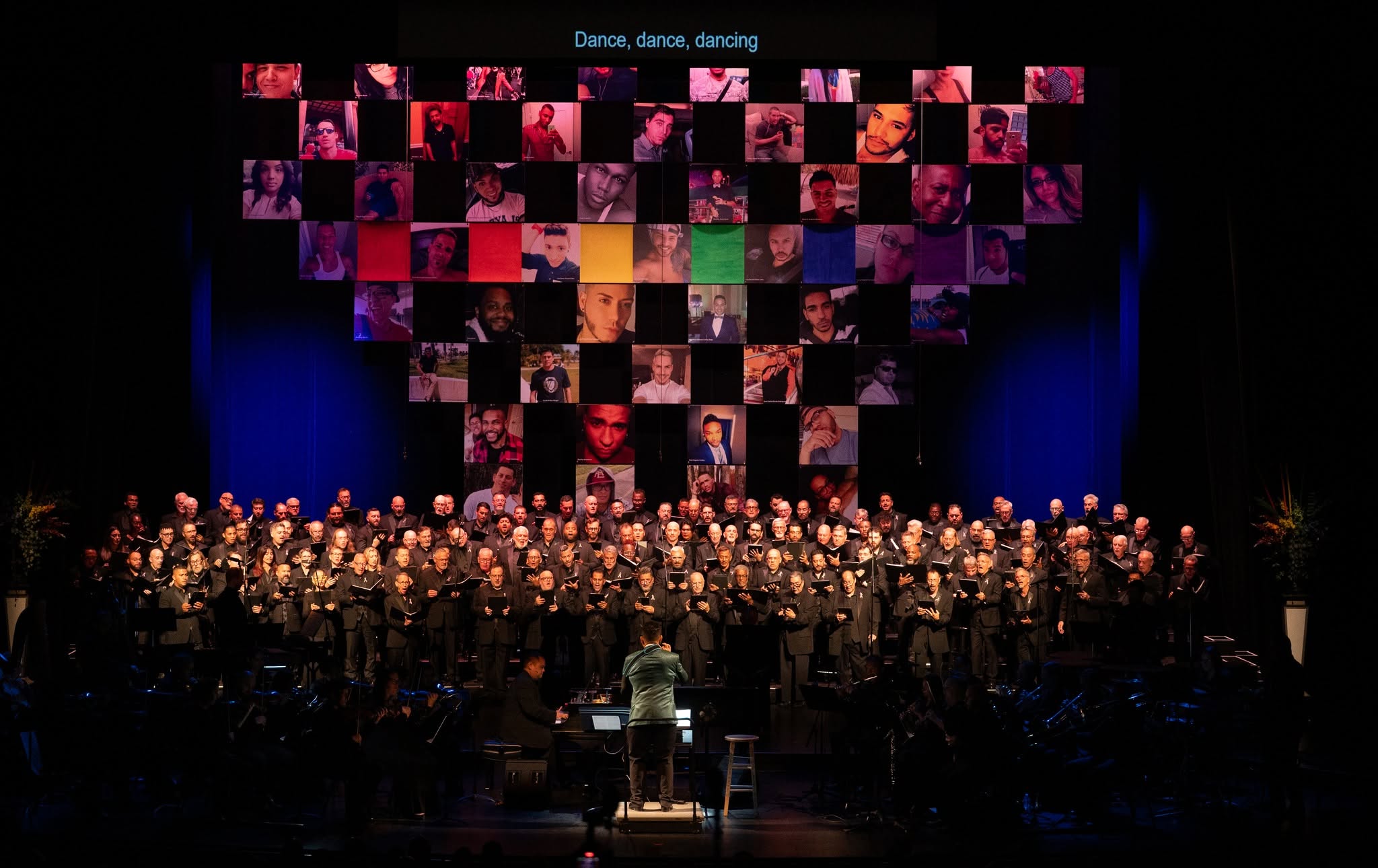A large choir in black attire performs on a dimly lit stage, with a mosaic wall of small portrait photos suspended above them and a conductor standing on a podium in front.