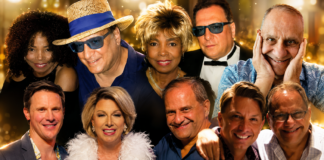 Group of ten adults smiling for a party photo, wearing colorful outfits against a golden bokeh backdrop.
