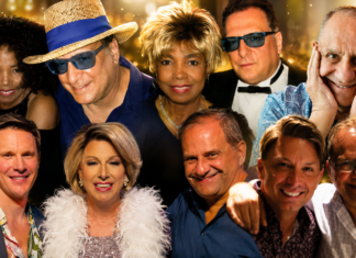 Group of ten adults smiling for a party photo, wearing colorful outfits against a golden bokeh backdrop.