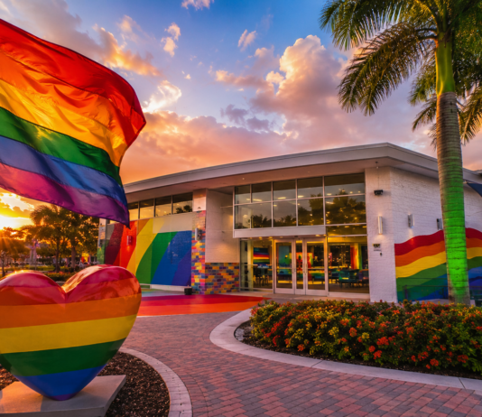 Rainbow flag waves above a rainbow-painted plaza in front of a modern building at sunset for a Pride-themed scene.