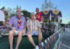People riding on a pride parade float wearing colorful outfits and rainbow accessories, with bead garlands on display behind them.