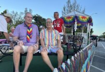People riding on a pride parade float wearing colorful outfits and rainbow accessories, with bead garlands on display behind them.