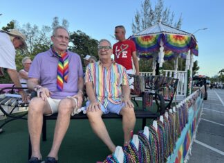 People riding on a pride parade float wearing colorful outfits and rainbow accessories, with bead garlands on display behind them.