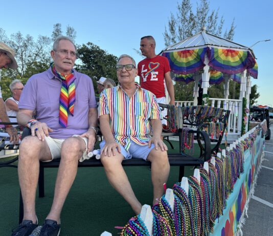 People riding on a pride parade float wearing colorful outfits and rainbow accessories, with bead garlands on display behind them.