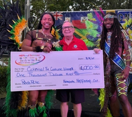 Paul Rolli: Ready to Lead Wilton Manors Into Its Next Chapter | Hotspots Magazine Hotspots Magazine Three people pose with a large ceremonial check at a carnival costume contest; two colorful feathered dancers flank an older man in a red polo shirt.