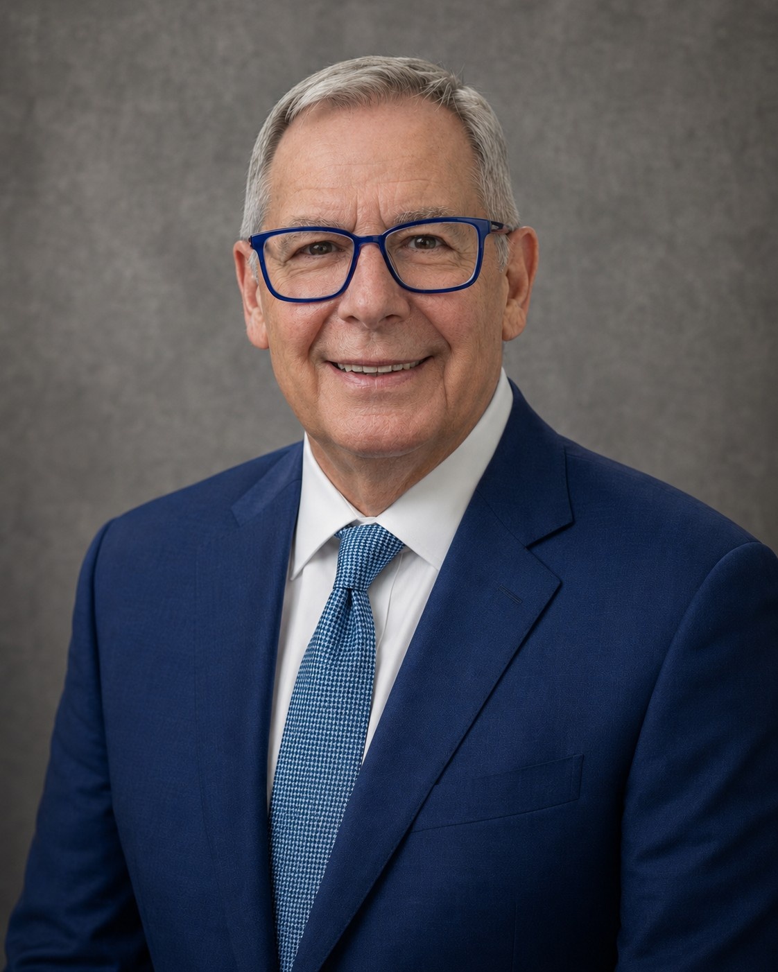 Paul Rolli: Ready to Lead Wilton Manors Into Its Next Chapter | Hotspots Magazine Hotspots Magazine Professional headshot of a smiling older man in a blue suit and patterned blue tie, wearing blue-framed glasses, with a gray backdrop.