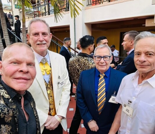 Four men in suits posing for a selfie at an outdoor event, name badges visible on each attendee.