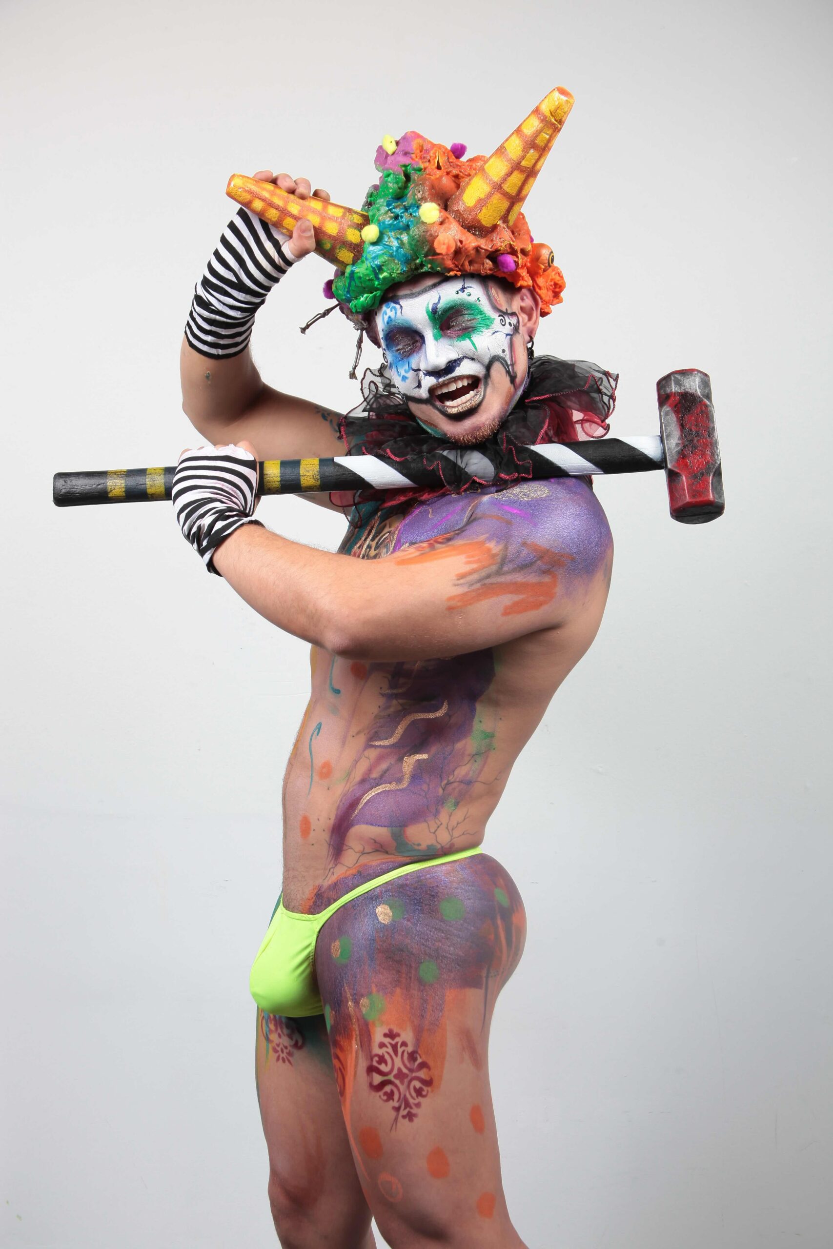 Person with vibrant body paint and clown makeup wears a colorful horned headdress and holds a striped hammer-like prop over the shoulder.