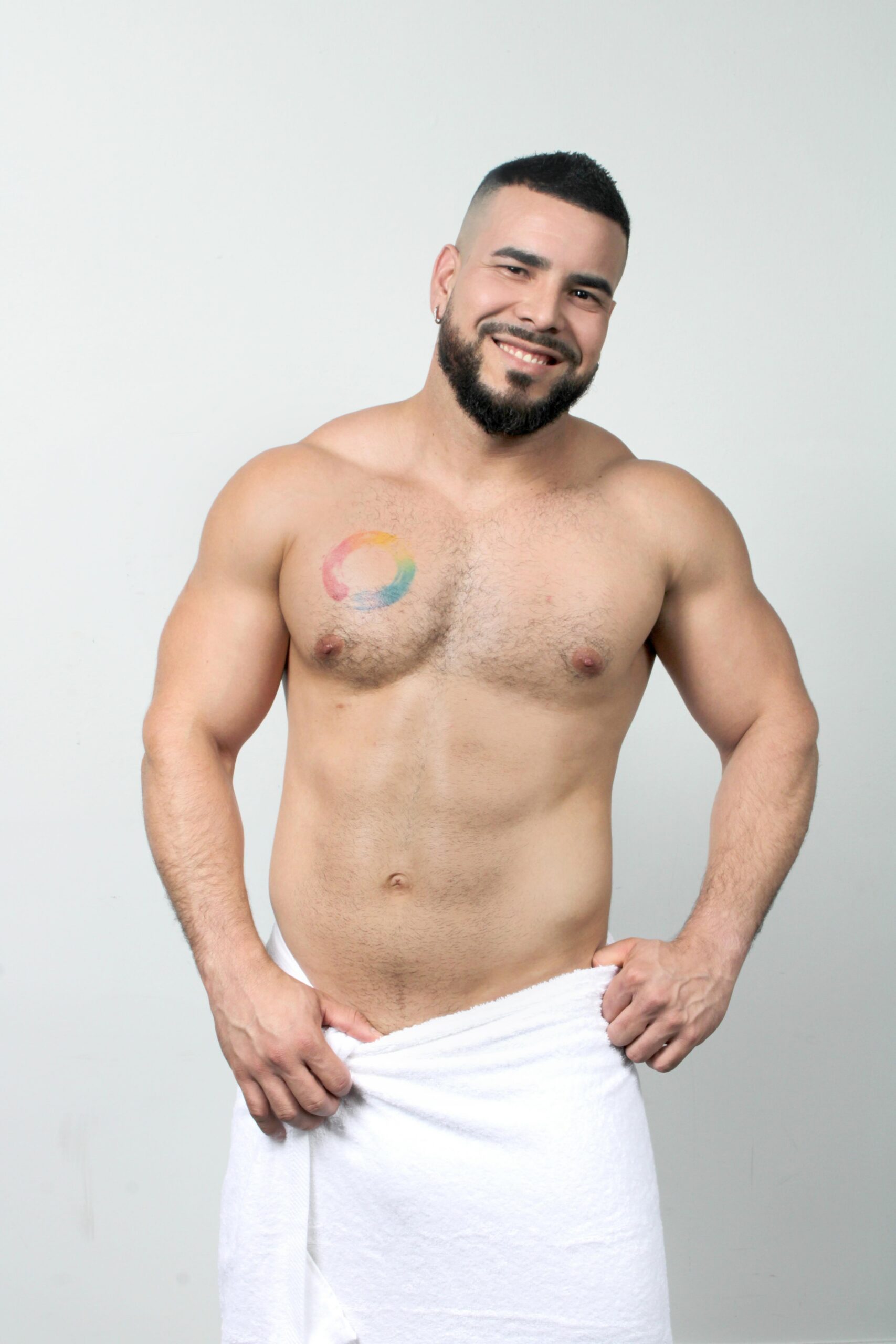 Smiling shirtless man with a rainbow circular tattoo on his left chest, wrapped in a white towel against a light background.