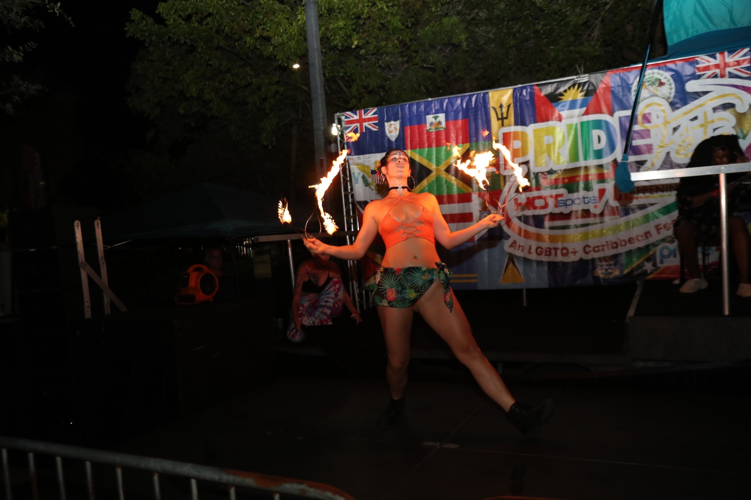Female fire performer spinning flaming wands on a stage at a Pride festival, wearing a coral top and tropical shorts with a colorful banner in the background.
