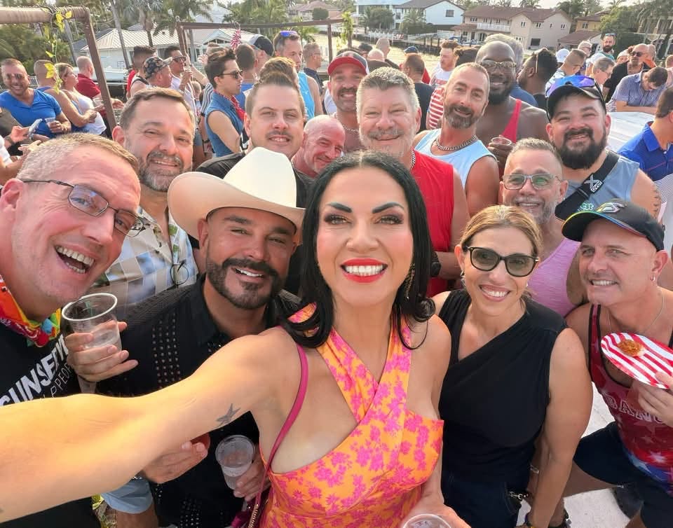 Group selfie at a lively outdoor party with many smiling attendees crowded together, woman in pink dress in center taking the photo.