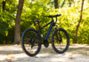 Black mountain bike parked on a sunlit path with green, leafy trees in the background.