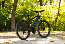 HOT Product: Huffy Eventis Mountain Ebike 27.5 inch Black mountain bike parked on a sunlit path with green, leafy trees in the background.