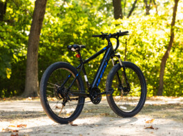 HOT Product: Huffy Eventis Mountain Ebike 27.5 inch Black mountain bike parked on a sunlit path with green, leafy trees in the background.