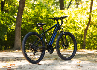 HOT Product: Huffy Eventis Mountain Ebike 27.5 inch Black mountain bike parked on a sunlit path with green, leafy trees in the background.