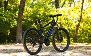 HOT Product: Huffy Eventis Mountain Ebike 27.5 inch Black mountain bike parked on a sunlit path with green, leafy trees in the background.