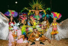 Group of carnival dancers in bright feathered costumes posing on a brick-paved ground at night, with a muscular dancer elevated above the crowd in the center.