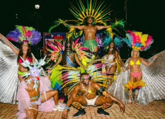 PrideFête: 5 Years Strong Group of carnival dancers in bright feathered costumes posing on a brick-paved ground at night, with a muscular dancer elevated above the crowd in the center.