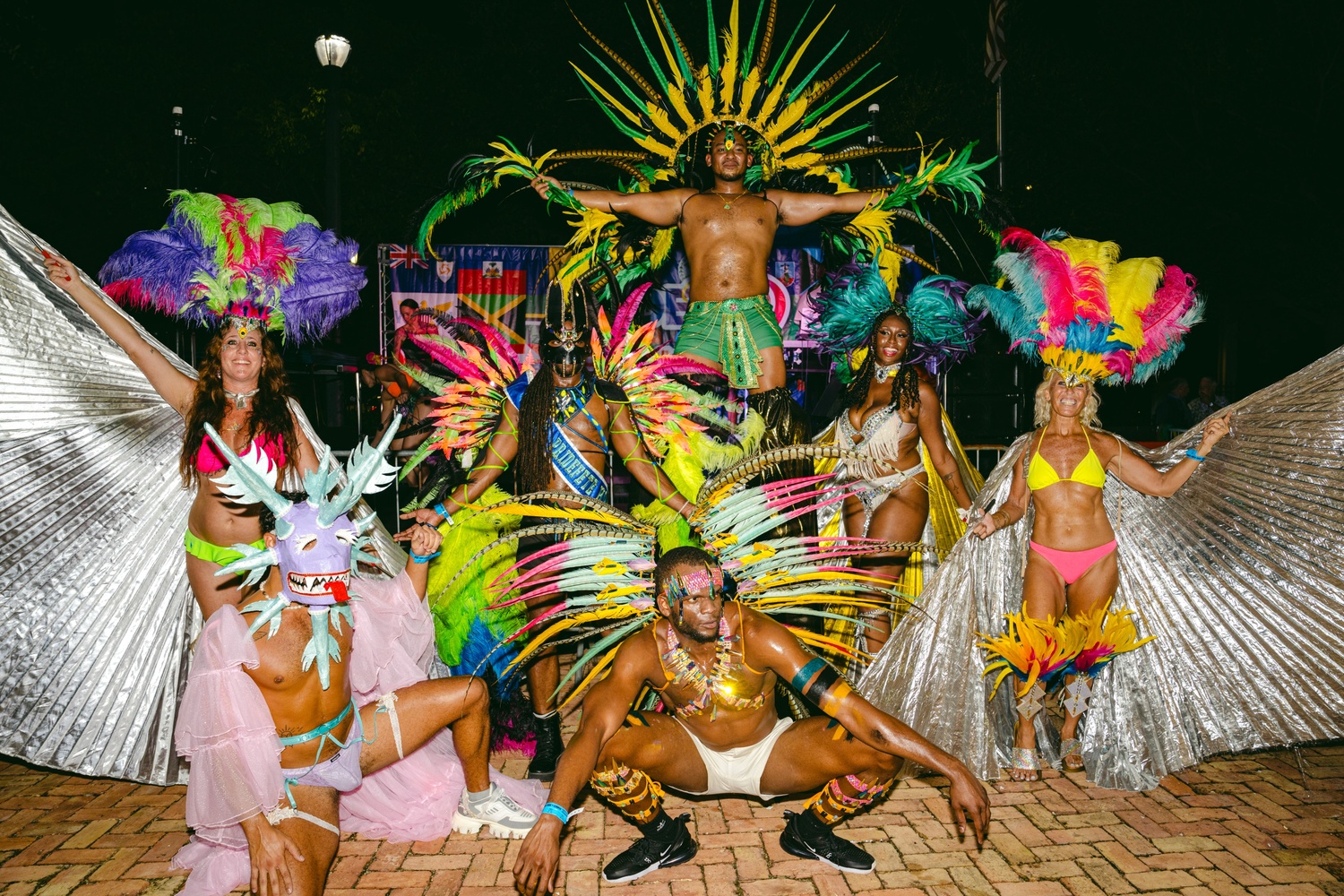 MEL00849 Group of carnival dancers in bright feathered costumes posing on a brick-paved ground at night, with a muscular dancer elevated above the crowd in the center.