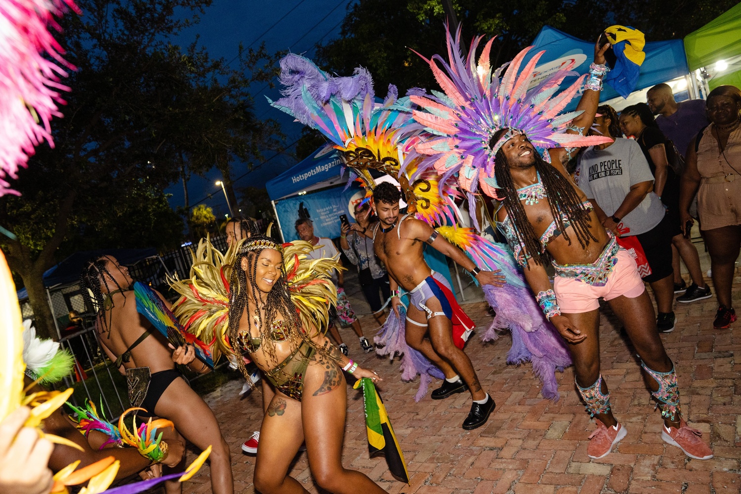 PrideFête: 5 Years Strong | Hotspots Magazine Hotspots Magazine Dancers in colorful feather costumes perform at a night street festival, with lights and onlookers in the background.