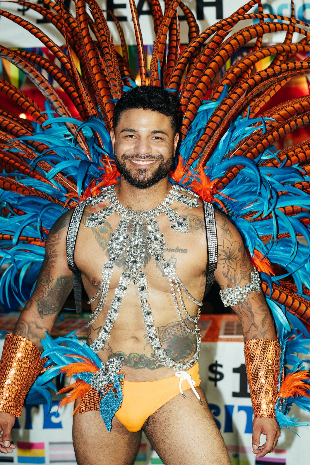 PrideFête: 5 Years Strong | Hotspots Magazine Hotspots Magazine Male carnival performer with a large orange-and-blue feather headdress, silver jeweled harness, orange swim trunks, and visible tattoos, smiling at the camera.