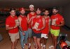 Six men in red shirts pose together in a bowling alley, each holding a brochure or flyer with a shirt logo visible in the background lanes.