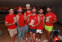 Six men in red shirts pose together in a bowling alley, each holding a brochure or flyer with a shirt logo visible in the background lanes.