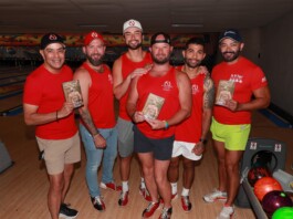 Six men in red shirts pose together in a bowling alley, each holding a brochure or flyer with a shirt logo visible in the background lanes.