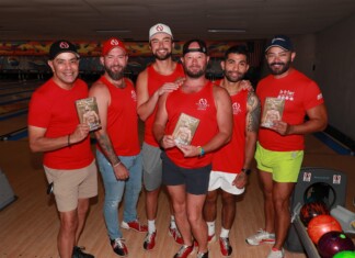 Out With Scott – By Scott Holland Six men in red shirts pose together in a bowling alley, each holding a brochure or flyer with a shirt logo visible in the background lanes.