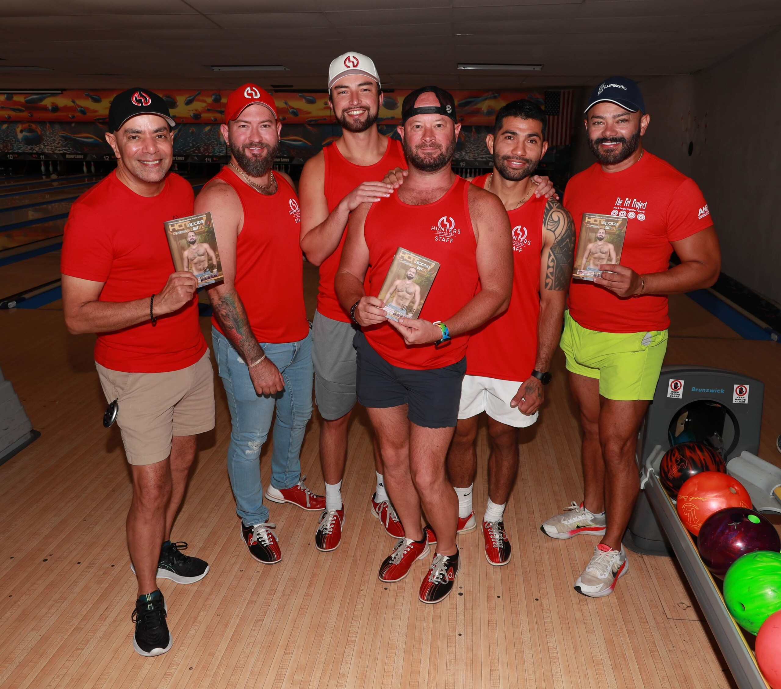 Pet Project Six men in red shirts pose together in a bowling alley, each holding a brochure or flyer with a shirt logo visible in the background lanes.