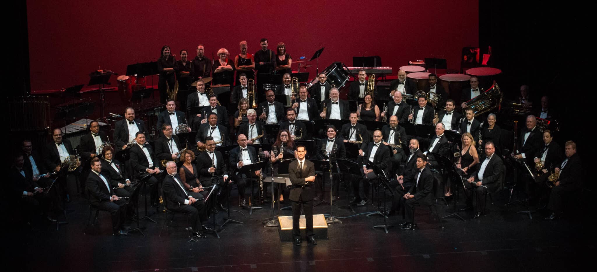 Conductor stands at a podium before a full symphony orchestra in formal black attire on a red-lit stage.