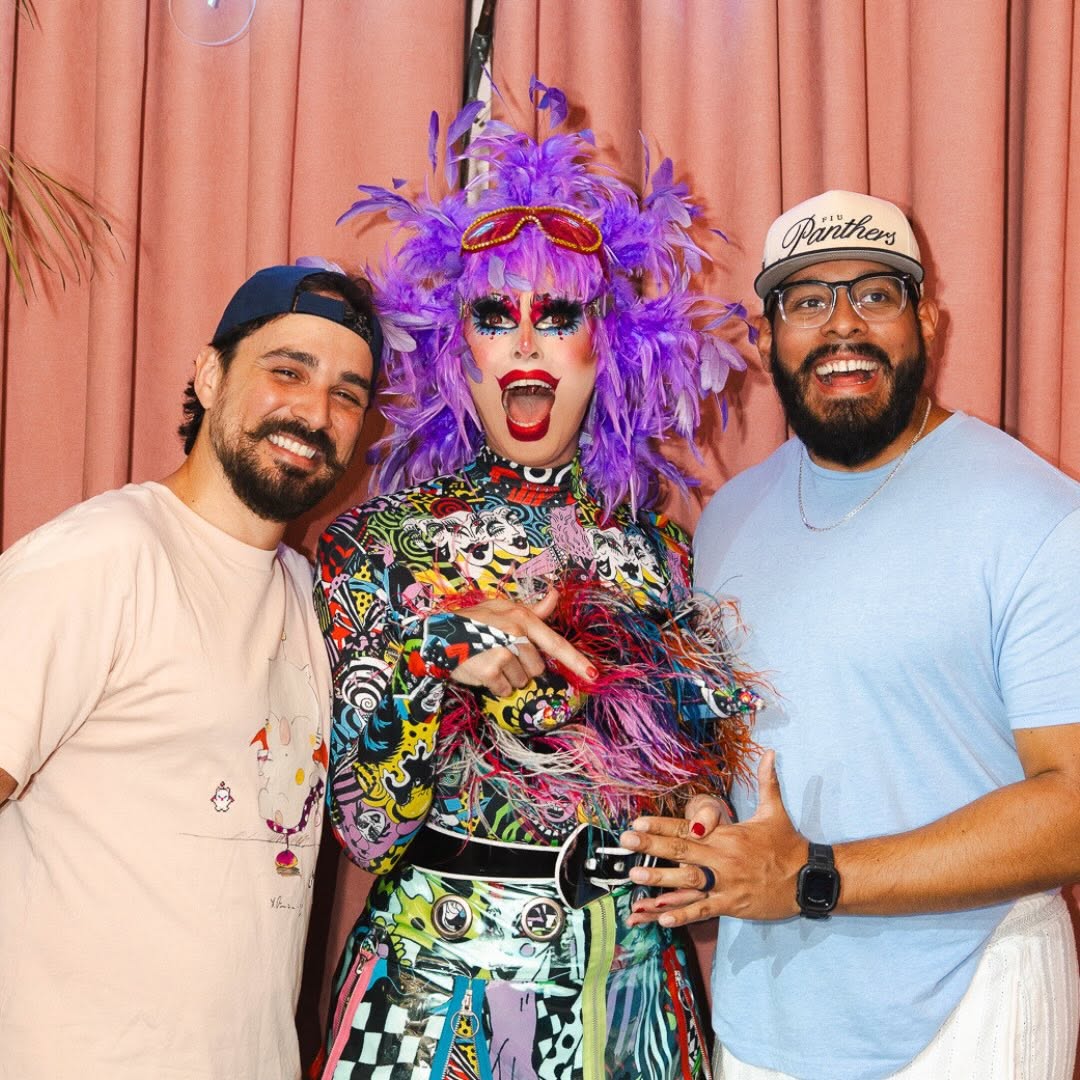 Drag queen wearing a colorful geometric costume and a purple feather wig posing with two smiling men in front of a pink curtain.