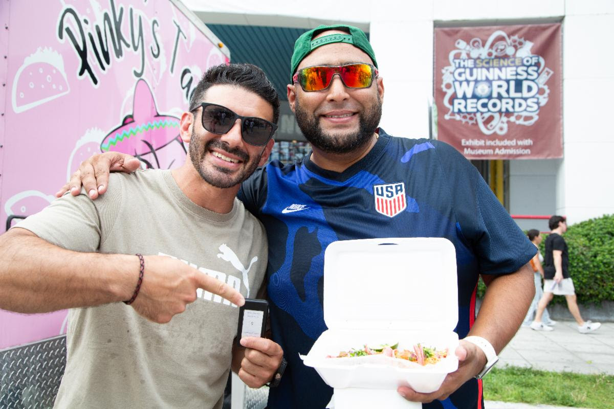 Out With Scott – By Scott Holland | Hotspots Magazine Hotspots Magazine Two smiling men pose together at an event; one holds a takeout container of food, the other points to a phone or device in his hand.