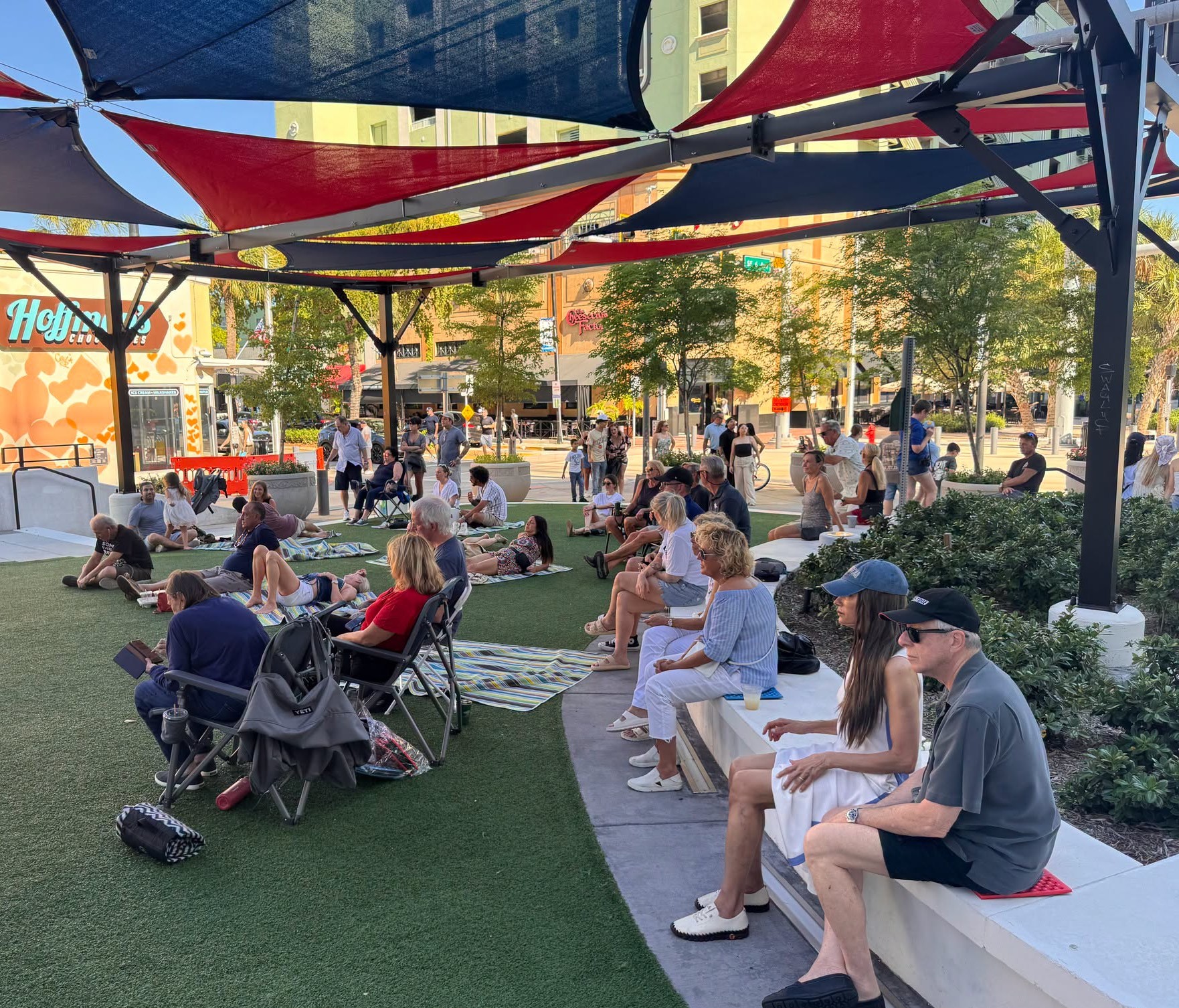 Crowd gathered at an outdoor event on a grassy plaza with colorful shade sails overhead and people sitting on blankets and chairs.