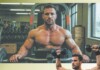 Bye Bye Man Boobs Muscular man shirtless using a dumbbell press machine in a gym, with another lifter and weights in the background.