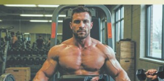 Bye Bye Man Boobs Muscular man shirtless using a dumbbell press machine in a gym, with another lifter and weights in the background.