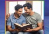 Two men sit closely together, smiling, as they read a book aloud to each other indoors.