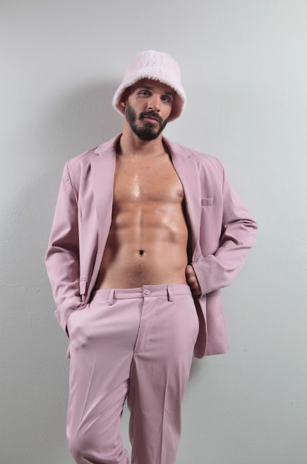 Male model in an open pink suit showing his bare chest and abs, wearing a pink bucket hat, against a light wall.