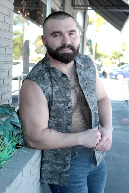 Bearded man with a sleeveless camo shirt leans against a brick planter outside a storefront, smiling slightly.