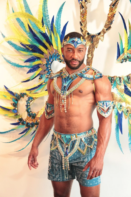 Male dancer in a blue-and-gold carnival costume with large multicolored feather wings and beaded accessories, posing against a light background.