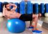Man performing an abdominal crunch on a blue stability ball in a gym with mirrors and equipment in the background.
