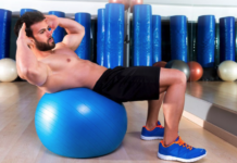 Focus on the Prize: How to Stay Focused on Your Fitness Goals Man performing an abdominal crunch on a blue stability ball in a gym with mirrors and equipment in the background.