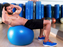 Focus on the Prize: How to Stay Focused on Your Fitness Goals Man performing an abdominal crunch on a blue stability ball in a gym with mirrors and equipment in the background.