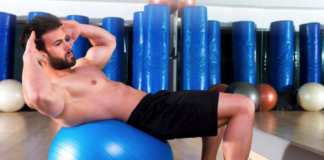 Man performing an abdominal crunch on a blue stability ball in a gym with mirrors and equipment in the background.